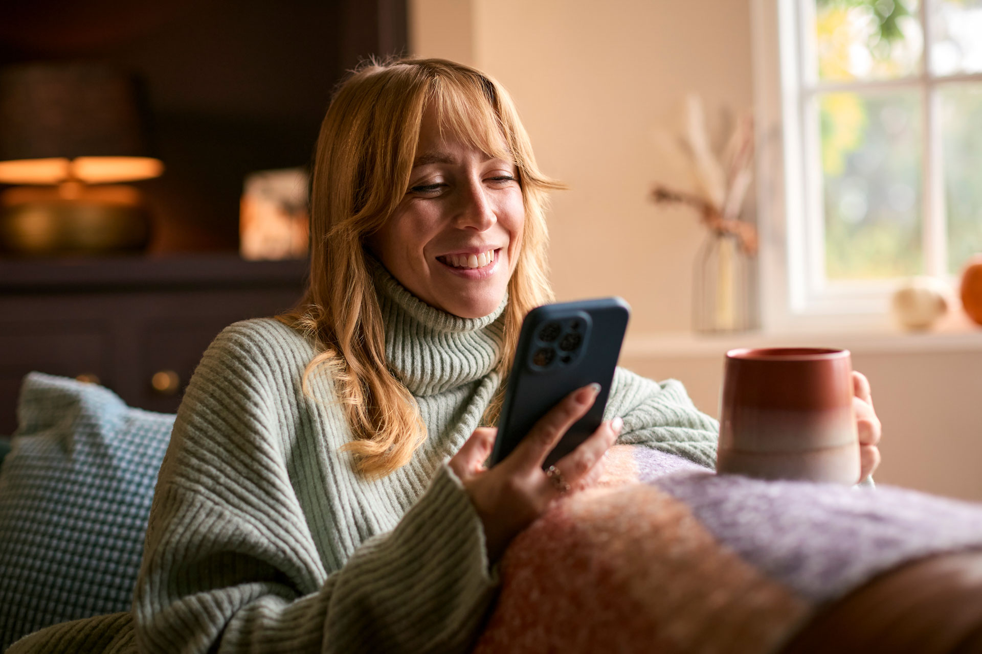A woman using the HBS Platform on her phone at home, while sat on her sofa holding a cup of tea and smiling