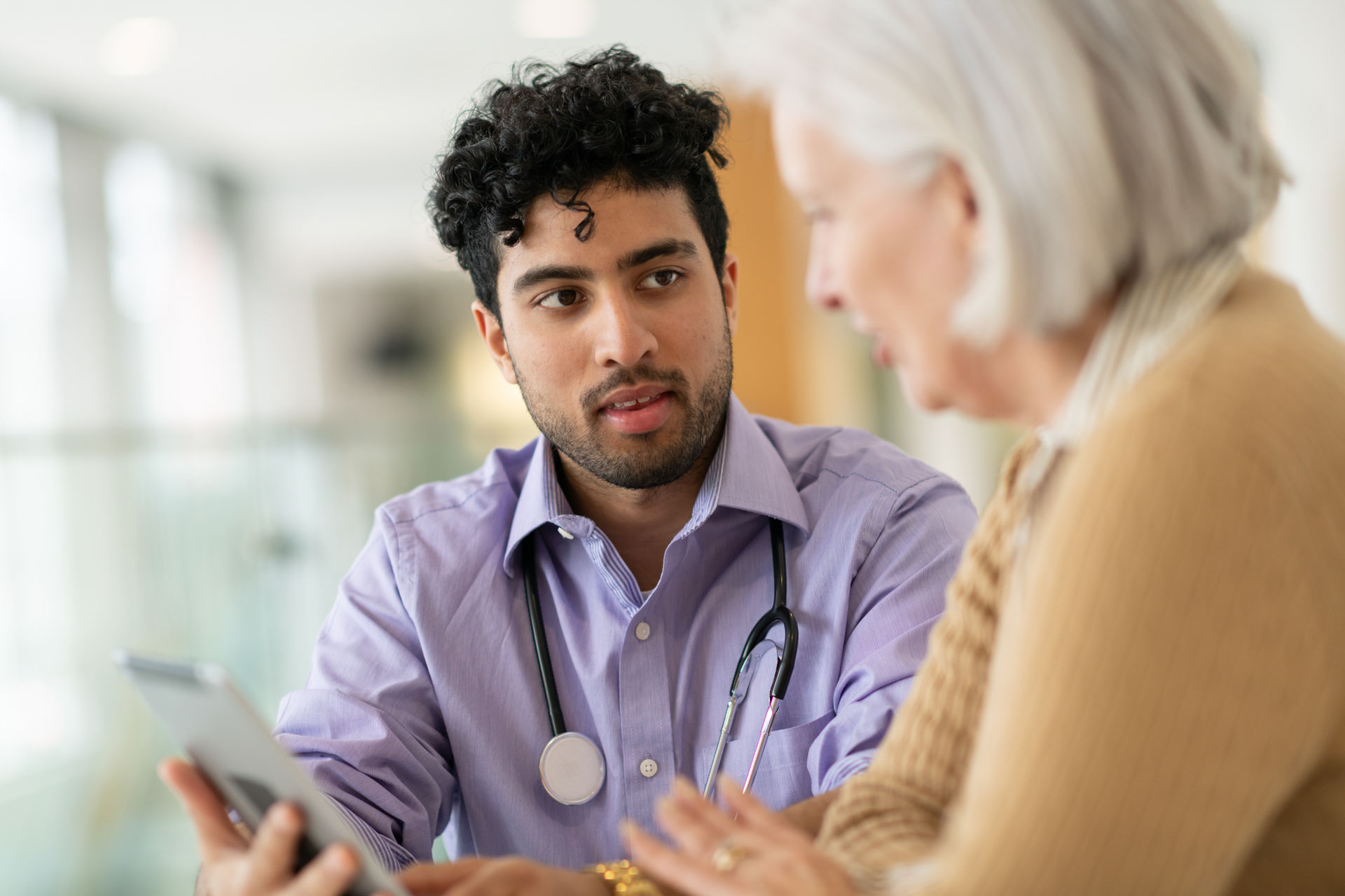 A multiracial doctor is doing a routine check-up with his elderly Caucasian female patient. They are seated at a table together and the doctor is holding a tablet computer. The two are reviewing test results and discussing a treatment plan. The patient is asking a question.