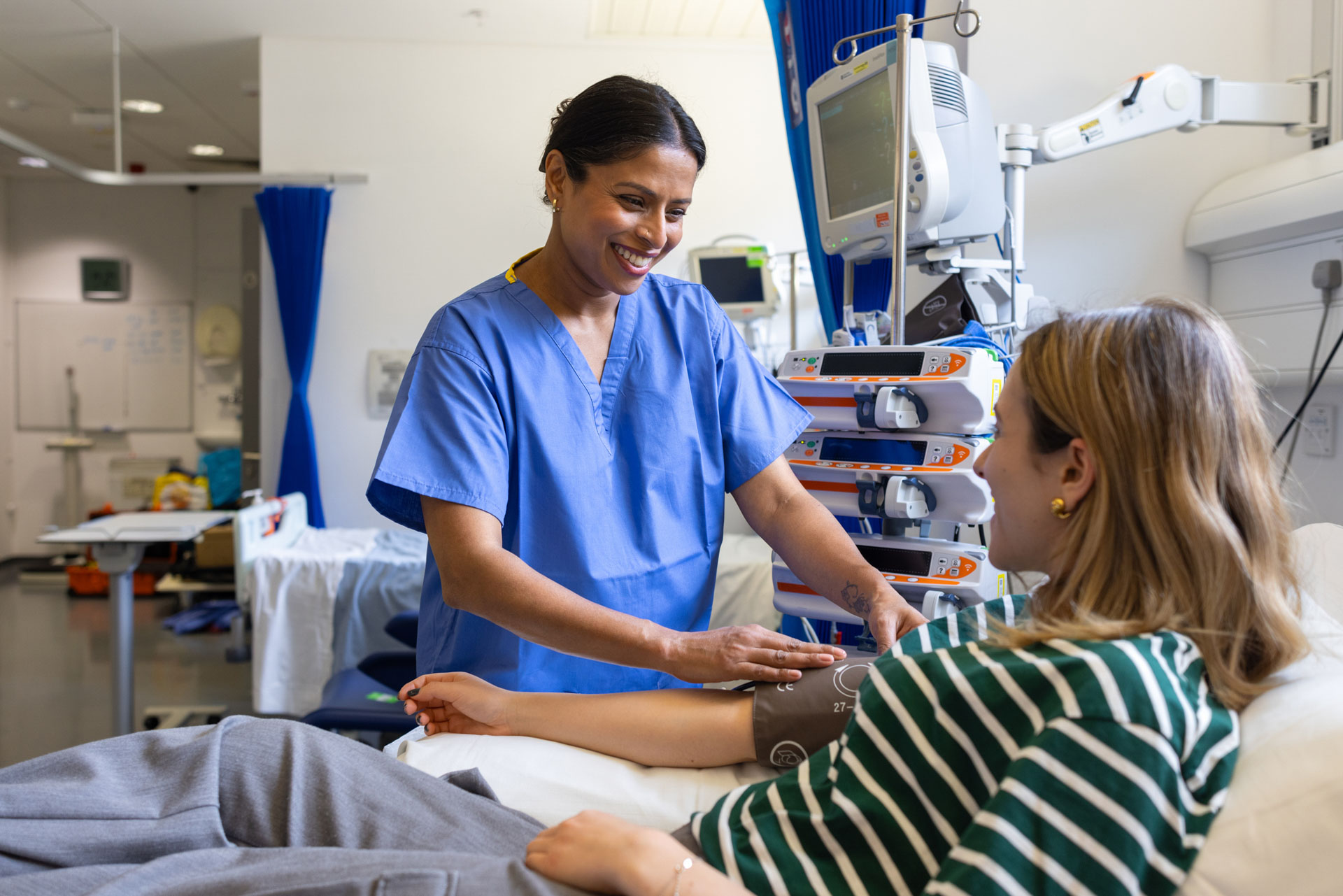 Medium shot of a young female in-patient sitting on a hospital bed, with a smiling nurse taking her blood pressure.