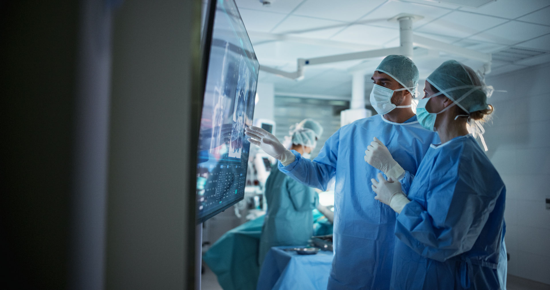 Two surgeons wearing hospital scrubs and face masks analysing scans on a digital monitor in an operating theatre