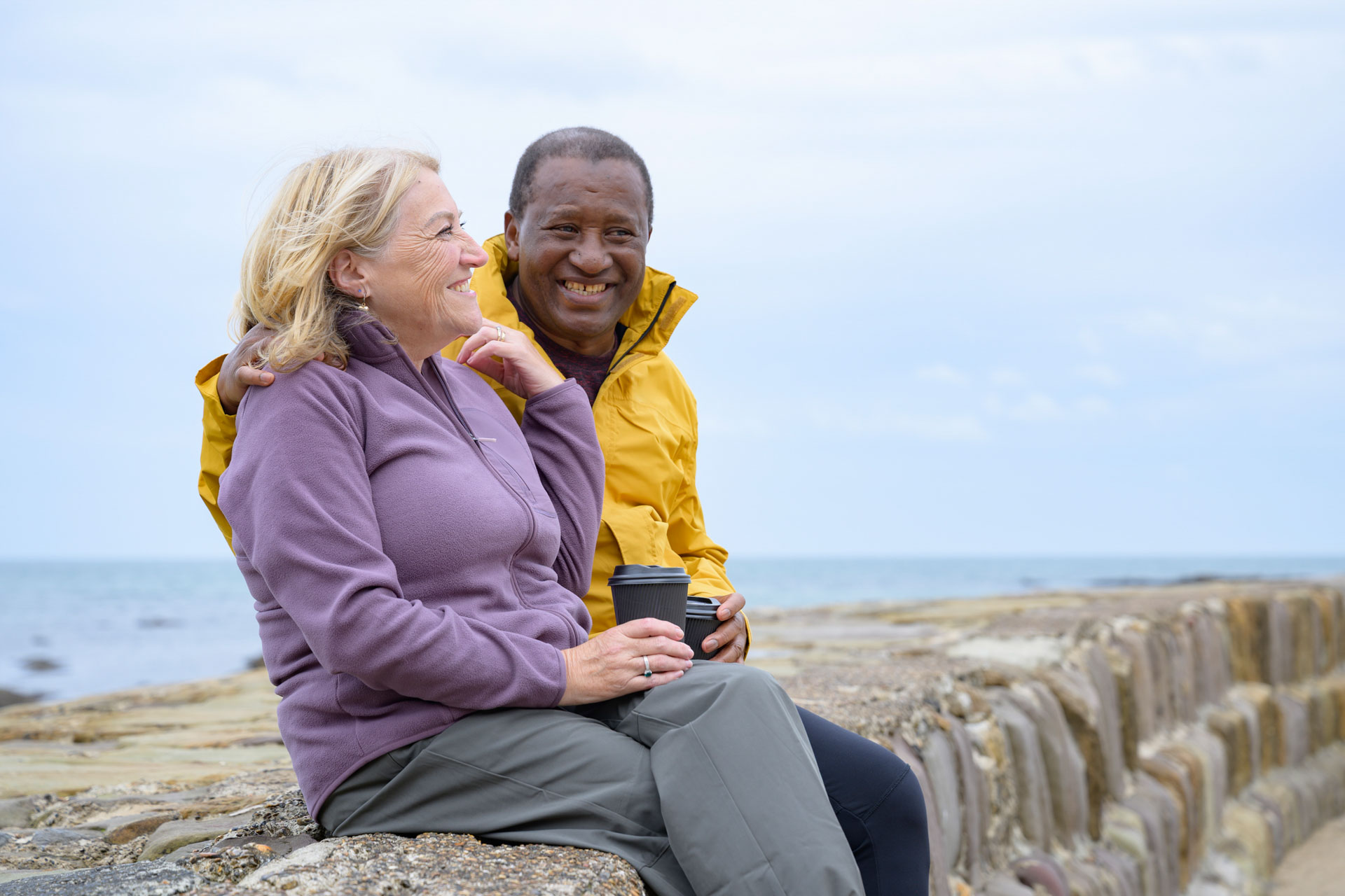 A happy older couple sitting on a sea wall enjoying a trip to the coast, laughing with each other.