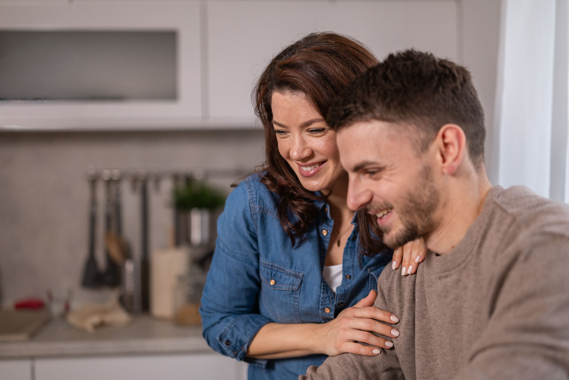 Happy couple embracing, smiling, and looking down at a laptop in a modern kitchen