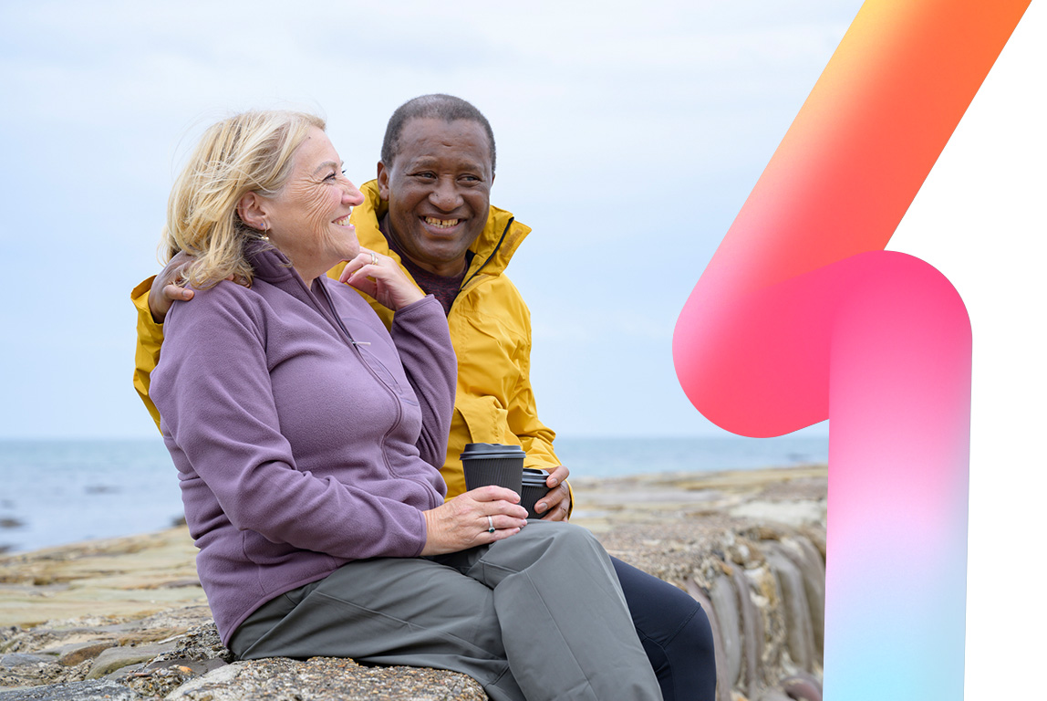 A happy older couple sitting on a sea wall enjoying a trip to the coast, laughing with each other.