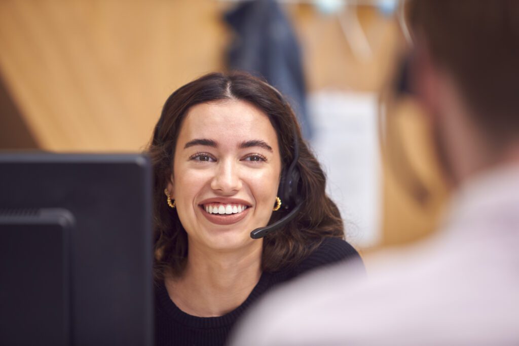 Businesswoman In Cubicle Wearing Headset Talking To Colleague In Busy Customer Services Centre